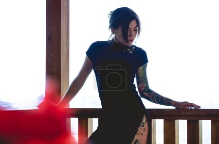 Téléchargez les photos : Élégance exotique : jeune femme brune sexy tatouée dans une robe chinoise bleue dans un pavillon en bois - en image libre de droit