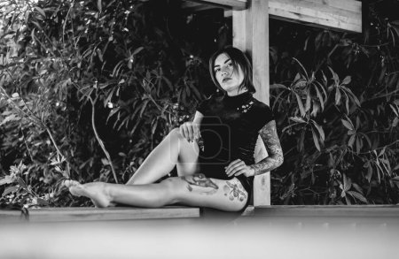 Téléchargez les photos : Élégance exotique : jeune femme brune sexy tatouée dans une robe chinoise bleue dans un pavillon en bois entouré de jungle (en noir et blanc) - en image libre de droit
