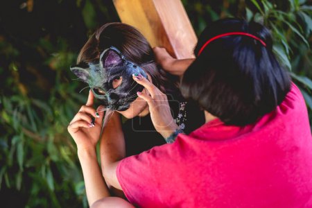 Téléchargez les photos : Jeune femme brune sexy tatouée dans une robe chinoise bleue avec un masque de chat en préparation dans un pavillon en bois entouré de jungle - en image libre de droit