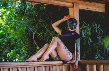 Téléchargez les photos : Séduction exotique : jeune femme brune sexy tatouée dans une robe chinoise bleue avec un masque de chat dans un pavillon en bois entouré de jungle - en image libre de droit