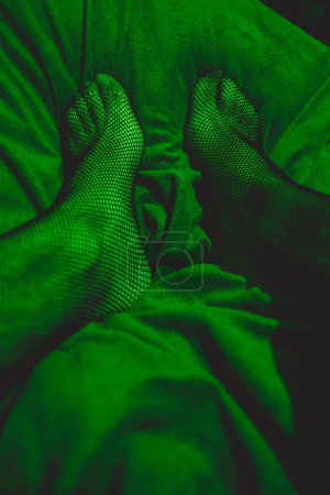 Téléchargez les photos : Détails des jambes et des pieds sexy en bas résille au lit sous la lumière verte séduisante et intime - en image libre de droit