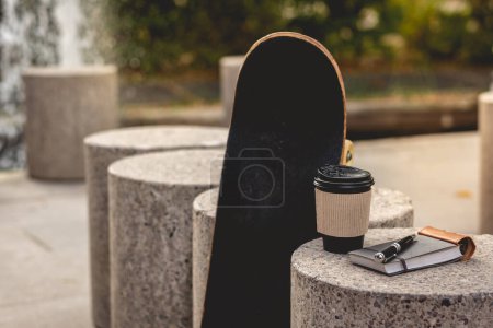 Foto de Ruptura de trabajo eficiente: cuaderno encuadernado en cuero, lápiz elegante, taza de café ecológica y monopatín en el soleado parque urbano - Imagen libre de derechos