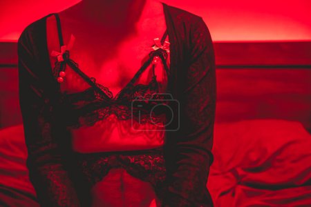Téléchargez les photos : Pliage des normes de genre : au lit avec un femboy non binaire en lingerie noire séduisante et lumière rouge passionnée - en image libre de droit