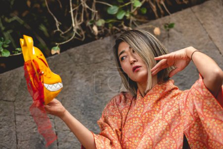 Téléchargez les photos : Tradition et beauté japonaises : femme aux cheveux blonds japonais en kimono de soie orange avec masque de kitsune, baignée de soleil matinal au parc japonais serein - en image libre de droit