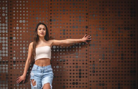 Téléchargez les photos : Ambiances urbaines : élégante fille latino en jeans et petit haut posant contre le mur de cuivre sous la lumière du soleil, avec un grand espace de copie - en image libre de droit