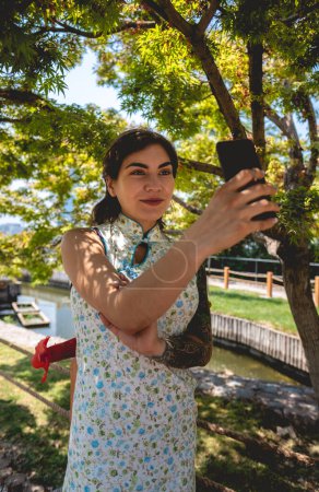 Téléchargez les photos : Technologie et beauté style japonais : fille brune avec des tatouages, robe asiatique, et téléphone faisant un selfie dans un jardin japonais - en image libre de droit