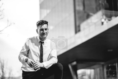 Téléchargez les photos : Jeune homme d'affaires professionnel, moderne et confiant avec chemise blanche et cravate noire tenant une tasse de café écologique en papier dans le parc urbain du quartier des affaires (en noir et blanc) - en image libre de droit