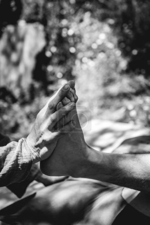 Toque curativo: detalle de un masajista realizando con la mano un masaje de pies y piernas en un entorno natural (en blanco y negro)