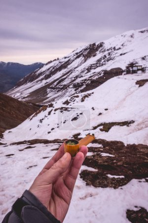 Téléchargez les photos : Fumer dans la neige : main tenant une pipe jaune avec de la marijuana, paysages de neige de montagne panoramiques - en image libre de droit