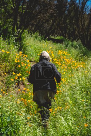 Téléchargez les photos : Aventure en montagne ensoleillée : un jeune photographe en tenue sombre explore une prairie ensoleillée parsemée de marguerites jaunes - en image libre de droit