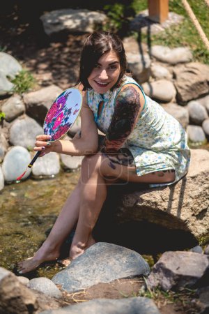 Multikulturelle Verschmelzung: Schöne Lateinerin in asiatischem Kleid, entspannt am Wasserstrom in einem japanischen Garten mit einem Papierfächer