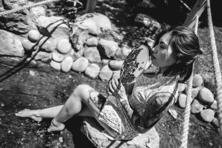 Multi cultural fusion: beautiful latin woman in an asian dress, relaxing by a water stream in a japanese garden with a paper fan (in black and white)