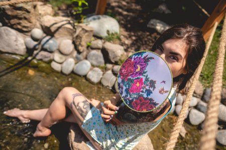 Multi cultural fusion: beautiful latin woman in an asian dress, relaxing by a water stream in a japanese garden with a paper fan