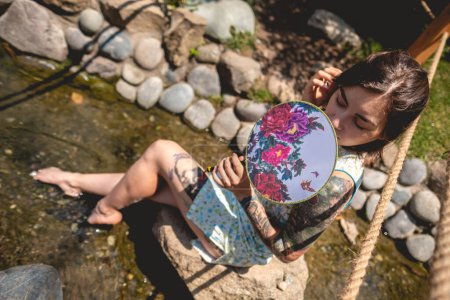 Multi cultural fusion: beautiful latin woman in an asian dress, relaxing by a water stream in a japanese garden with a paper fan