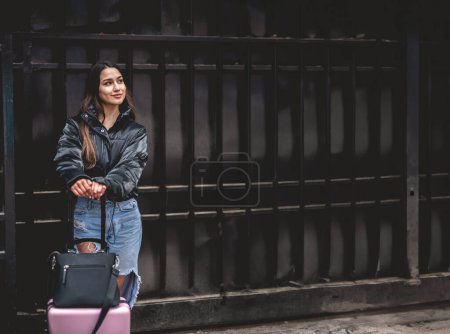 Téléchargez les photos : Voyage élégant et exploration urbaine : jeune brune latina vibrante avec look mode et valise rose explorant la ville au centre-ville - en image libre de droit