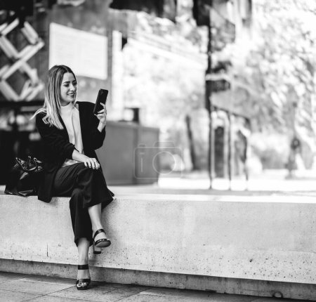 Téléchargez les photos : Pause café dans un lieu public de la ville : belle et détendue blonde latina cadre en utilisant le téléphone (en noir et blanc) - en image libre de droit