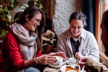 Photo for Two happy female friends enjoy coffee time in winter time indoors. They wear casual clothes like jacket, jeans, or scarf. Latte and cakes dessert on the wooden table. Concept: friendship - Royalty Free Image