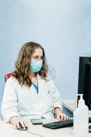 Photo for A young student female doctor wearing a medical gown check at her computer desk in a hospital. Professional healthcare provider offering medical advice - Royalty Free Image