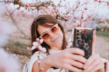 Téléchargez les photos : Photo horizontale d'une jolie femme souriante avec des lunettes de soleil et un pull hipster prenant un portrait selfie dans un champ de cerisiers en fleurs au printemps. Coucher de soleil et lumière douce. Concept de médias sociaux - en image libre de droit