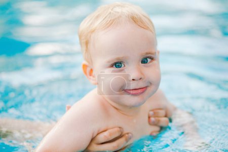 Photo for Portrait of small red-haired boy bathes in an open-air swimming pool in the garden with hand support, baby plays in water, family summer leisure - Royalty Free Image