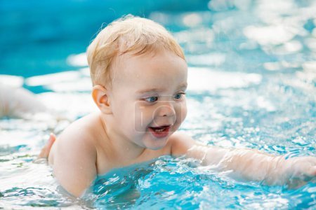 Retrato de niño de pelo rojo pequeño se baña en una piscina al aire libre en el jardín con soporte para las manos, el bebé juega en el agua, ocio familiar de verano