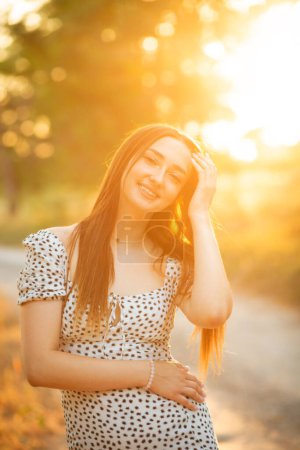 Téléchargez les photos : Mignonne fille souriante éclairée par la lumière du soleil chaud sur un chemin qui vous regarde, jeune femme romantique dans une robe marchant au coucher du soleil dans une forêt plantation, détente sur la nature - en image libre de droit