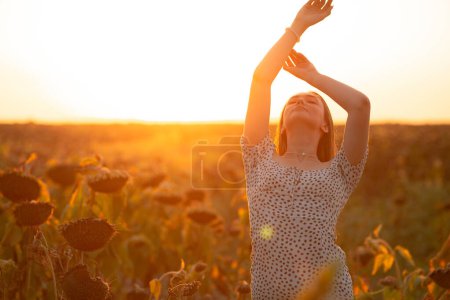 Téléchargez les photos : Jeune femme dans la lumière du soleil du soir dans un champ de tournesol au coucher du soleil d'été, fille tenant ses mains dans l'air, détente sur le concept de la nature - en image libre de droit