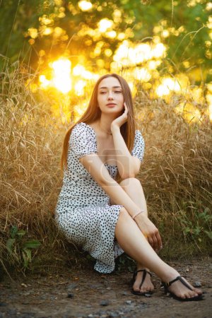 Téléchargez les photos : Portrait de jeune femme romantique dans une robe assise sur le bord de la route de la forêt au coucher du soleil, promenade d'été, concept de détente sur la nature - en image libre de droit