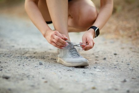 Téléchargez les photos : Jeune femme attachant des lacets de chaussures de course pour la course d'été dans le parc forestier. Le coureur se prépare pour la course. Jogging fille exercice, mode de vie sain concept - en image libre de droit