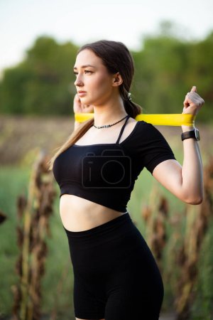 Téléchargez les photos : Jeune belle femme travaillant avec des élastiques de fitness dans le domaine du tournesol sec, mains d'entraînement de fille, concept de sport - en image libre de droit