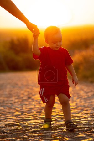 Téléchargez les photos : Silhouette de petit garçon tenant la main des parents sur une route en pierre, mère avec son fils marchant à l'extérieur au coucher du soleil - en image libre de droit