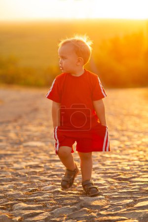 Téléchargez les photos : Mignon petit blond garçon silhouette marche sur la route de pierre dans la lumière du soleil, un enfant à l'extérieur au coucher du soleil, concept d'enfance - en image libre de droit