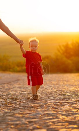 Téléchargez les photos : Silhouette de petit garçon tenant la main des parents sur une route en pierre, mère avec son fils marchant à l'extérieur au coucher du soleil - en image libre de droit