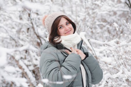 Téléchargez les photos : Portrait de jolie jeune femme en bonnet tricoté et foulard chaud parmi les arbres recouverts de neige, fille heureuse marchant sur la forêt d'hiver jouissant d'une belle nature - en image libre de droit