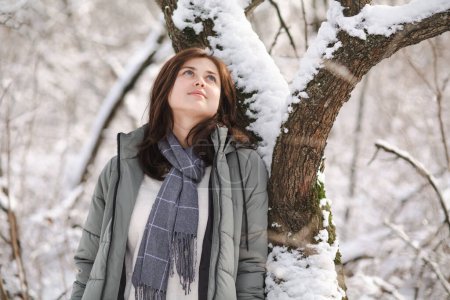 Téléchargez les photos : Jolie jeune femme en vêtements d'hiver se pencher sur l'arbre couvert de neige, fille marchant dans la forêt enneigée profiter de la belle nature - en image libre de droit