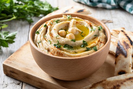 Téléchargez les photos : Hummus de pois chiches dans un bol en bois garni de persil, paprika et huile d'olive sur table en bois - en image libre de droit