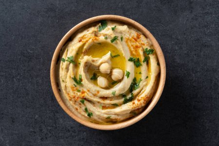 Téléchargez les photos : Hummus de pois chiches dans un bol en bois garni de persil, paprika et huile d'olive sur fond d'ardoise noire. Vue du dessus - en image libre de droit