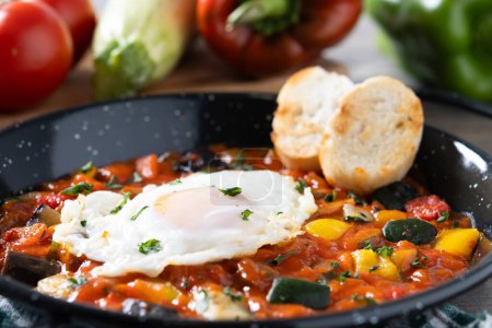 Gemüse Pisto Manchego mit Tomaten, Zucchini, Paprika, Zwiebeln, Auberginen und Ei, serviert in Pfanne auf Holztisch. Nahaufnahme