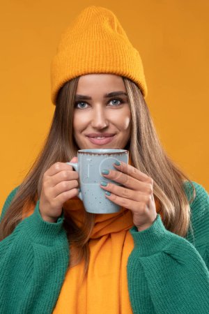 Photo for Young woman wearing a winter hat and scarf with a mug on an orange background. - Royalty Free Image