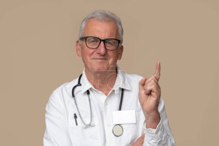 Foto de Médico mayor en gafas graduadas con estetoscopio y uniforme blanco sobre fondo marrón claro. - Imagen libre de derechos