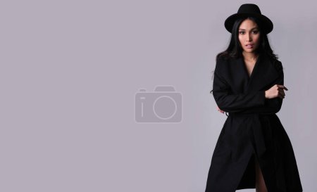 Téléchargez les photos : À la mode brune asiatique ethnicité dans élégant manteau noir et chapeau od fond gris. - en image libre de droit