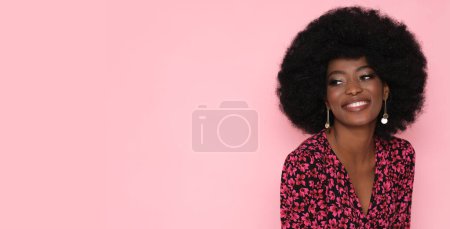 Foto de Bonita morena con peinado afro en vestido floral rojo sobre fondo rosa aislado. - Imagen libre de derechos