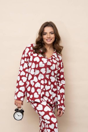 Foto de Atractiva morena en pijama de corazón rojo y blanco con un despertador en la mano sobre un fondo marrón claro en el estudio. - Imagen libre de derechos