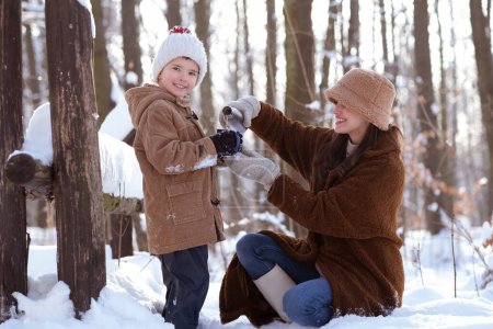 Foto de Mamá e hijo tienen un descanso de té de invierno en la hermosa zona de bosque nevado. - Imagen libre de derechos