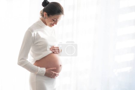 Photo for Beautiful brunette with a pregnant belly against the background of white window curtains with space for text. - Royalty Free Image