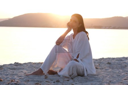 Téléchargez les photos : Séance photo romantique de beauté brune en vêtements à la mode sur la plage pendant le coucher du soleil. - en image libre de droit