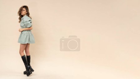 Téléchargez les photos : Séance photo sensuelle de beauté brune en robe de menthe pastel et bottes sur fond crémeux. - en image libre de droit