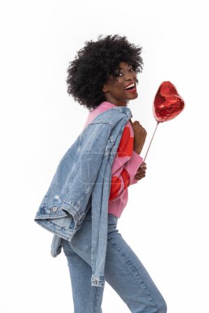 Photo for Happy black woman with afro hair and denim outfit is holding ballon heart on isolated white background. - Royalty Free Image