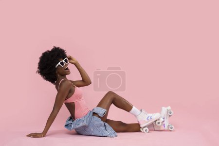 Téléchargez les photos : Heureuse femme noire avec patins à roulettes sur fond rose pastel. - en image libre de droit