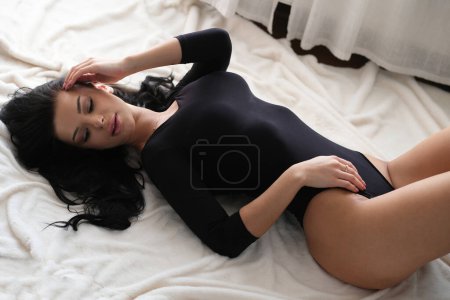 Téléchargez les photos : Une belle brune sexy en body noir serré repose sur une couverture blanche. - en image libre de droit